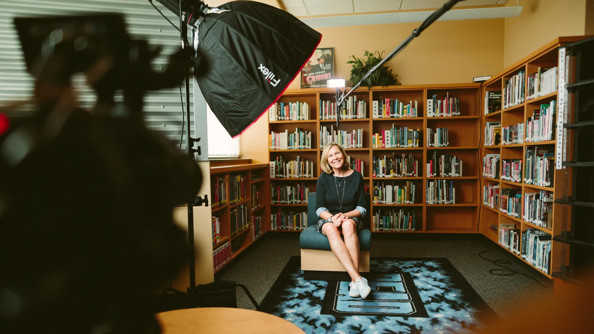 Woman sitting in front of bookcases in a comfortable chair, with lights, cameras, and microphones surrounding her as the interview subject.